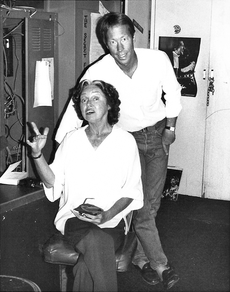 A woman sits on a chair"," gesturing with her hand"," while a man stands behind her"," both casually dressed in a room with posters.