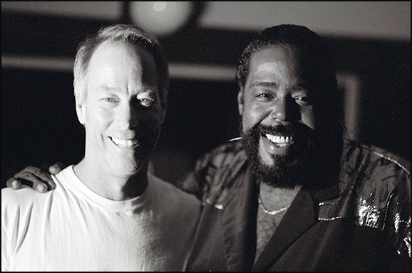 Two smiling men standing close together"," one man with light hair in a plain T-shirt and the other a man with a beard wearing a dark patterned shirt.