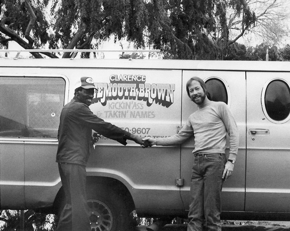 Two men standing on the passenger side of a 1970s van shaking hands with arms extended as if to showcase the decal on the side of the musician's tour vehicle.