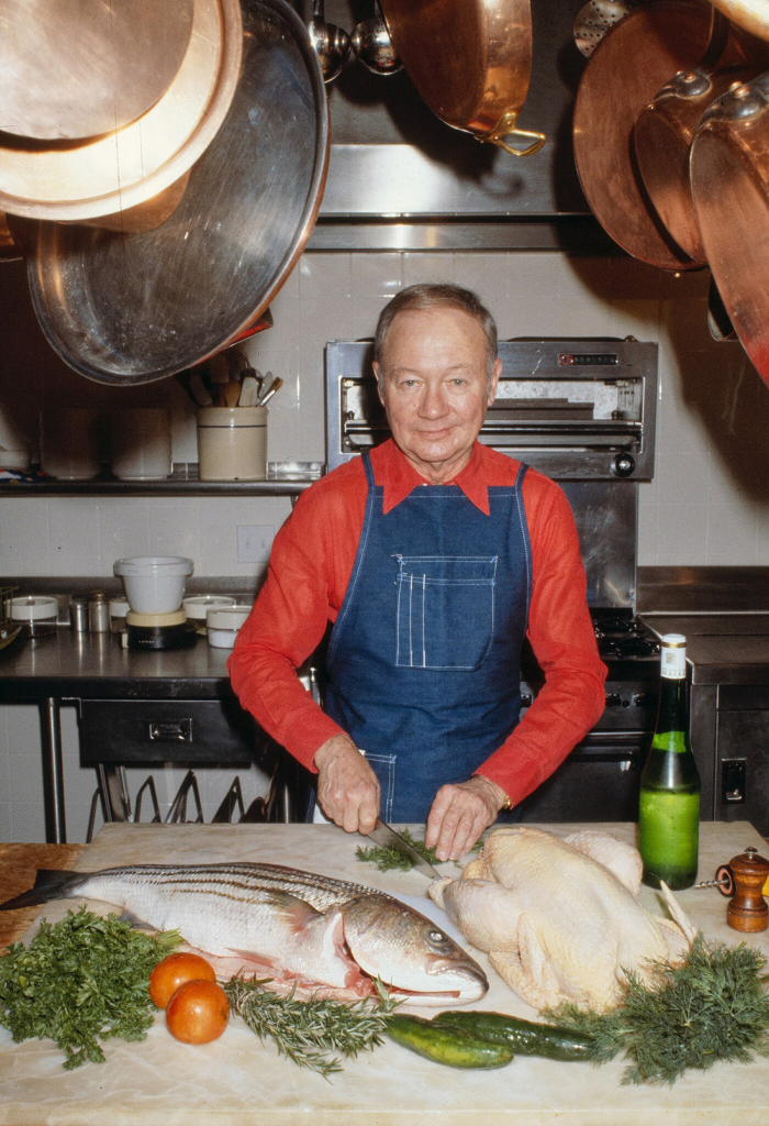 A middle-aged man wearing a red long-sleeved shirt and denim apron stands at a counter chopping herbs. In front of him are ingredients for cooking, including a fish and turkey. Copper pots hang overhead.