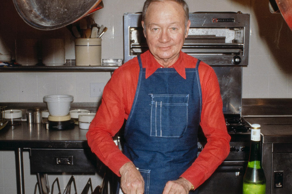 A middle-aged man wearing a red long-sleeved shirt and denim apron stands at a counter chopping herbs. In front of him are ingredients for cooking, including a fish and turkey. Copper pots hang overhead.