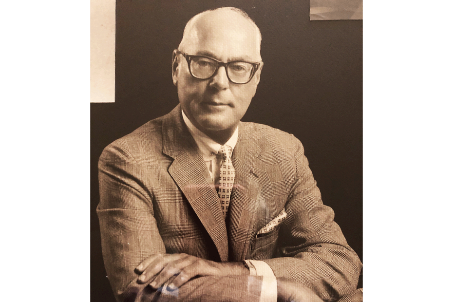 A man wearing glasses, an elegant suit, and tie poses with his arms crossed looking directly at the camera
