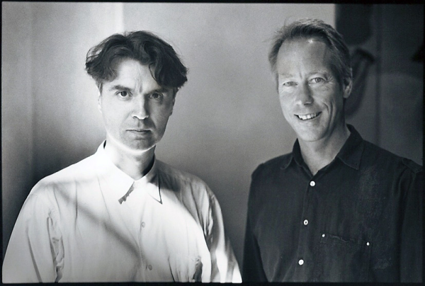 Two men standing together. The man on the left wears a light color shirt and gazes directly without smiling. The man on the right smiles and wears a dark colored shirt.