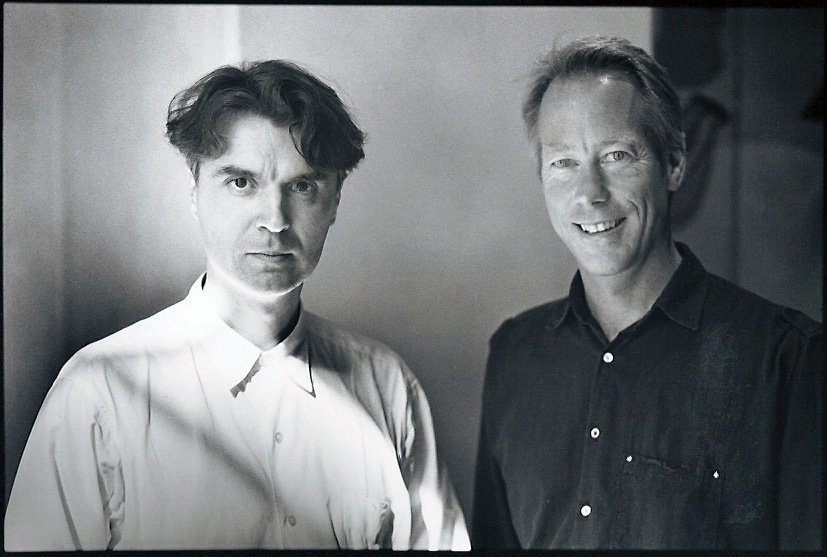 Two men standing together. The man on the left wears a light color shirt and gazes directly without smiling. The man on the right smiles and wears a dark colored shirt.