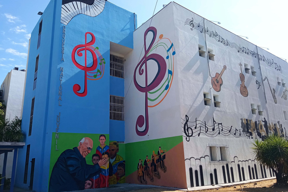 Building exterior in blue and white, with musical notes, piano keys, and other instruments painted on the walls. A mural on the bottom left shows an elderly man conducting a children's choir and musicians.