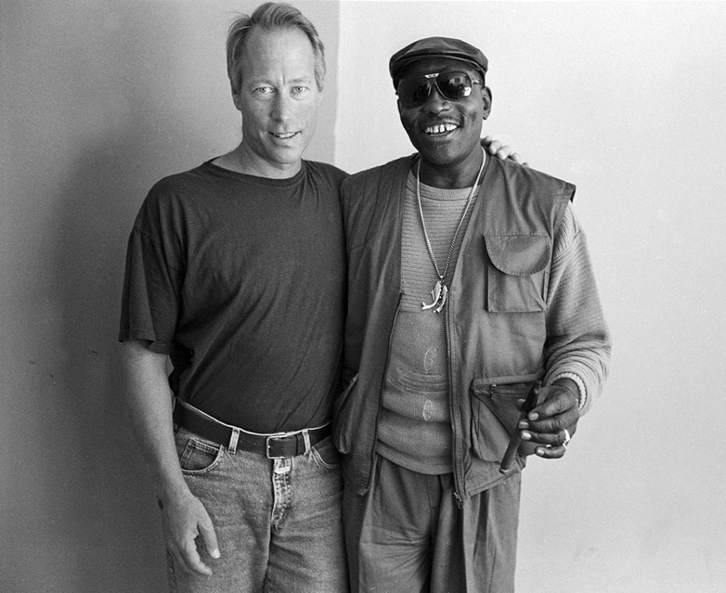 Two smiling men stand together. The man on the left wears a plain dark T-shirt and rests his arm around the shoulder of the man on the right, who wears a cap, sunglasses, vest over a sweater, a long necklace, and holds a cigar.