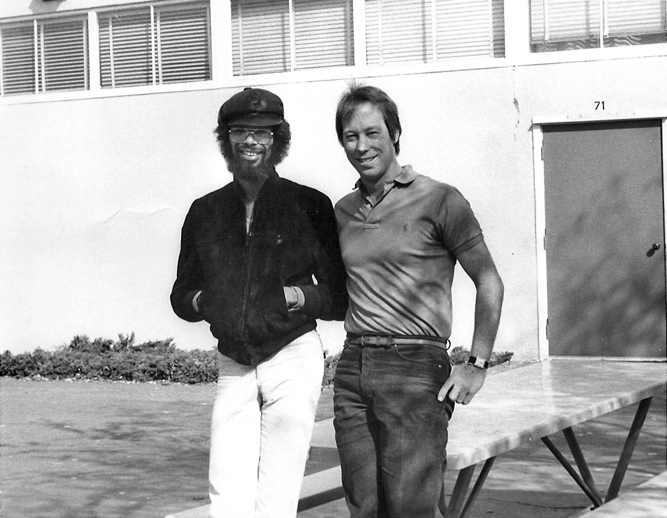 Two smiling men stand together outside in front of a picnic table and bench. The bearded man on the left wears a cap, sunglasses, and a dark colored jacket. The man on the right wears a polo shirt and a watch.