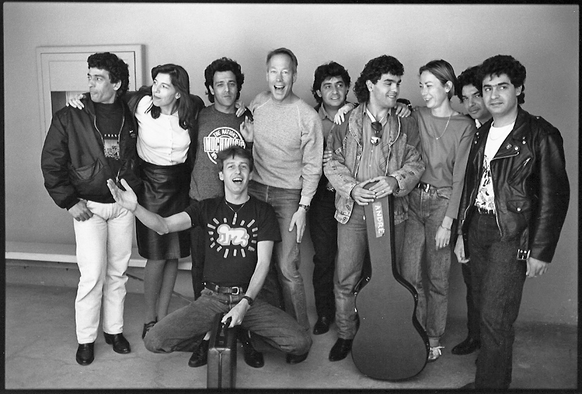 A group of ten people in casual clothing, smiling and posing together with arms around each other, one holding a guitar case.