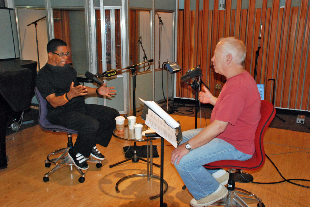 Two seated men in conversation inside a recording studio with microphones in front of them. The man on the left wears glasses and all-black clothing, gesturing with arms out and palms open. The man on the right wears jeans and a red T-shirt, with his left arm on his knee and right arm partially raised.