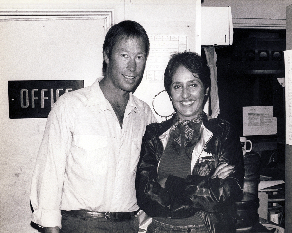 A woman with short hair wearing a scarf and jacket stands with her arms crossed next to a man in a light colored shirt to her left, both smiling, in an office space.