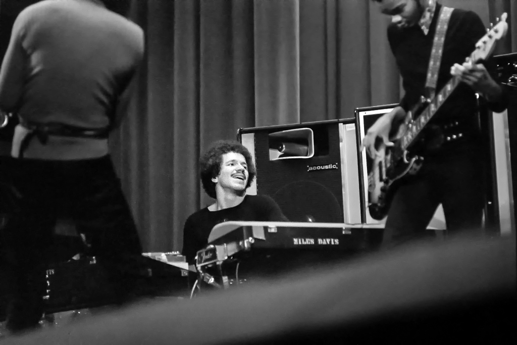 A young man with a mustache sits behind a keyboard while a man stands with his back to the camera at his left and a man plays the guitar on the right