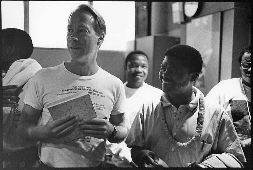 A group of people, including a man holding a folder and wearing a t-shirt, stand together indoors, smiling and interacting in a candid moment.