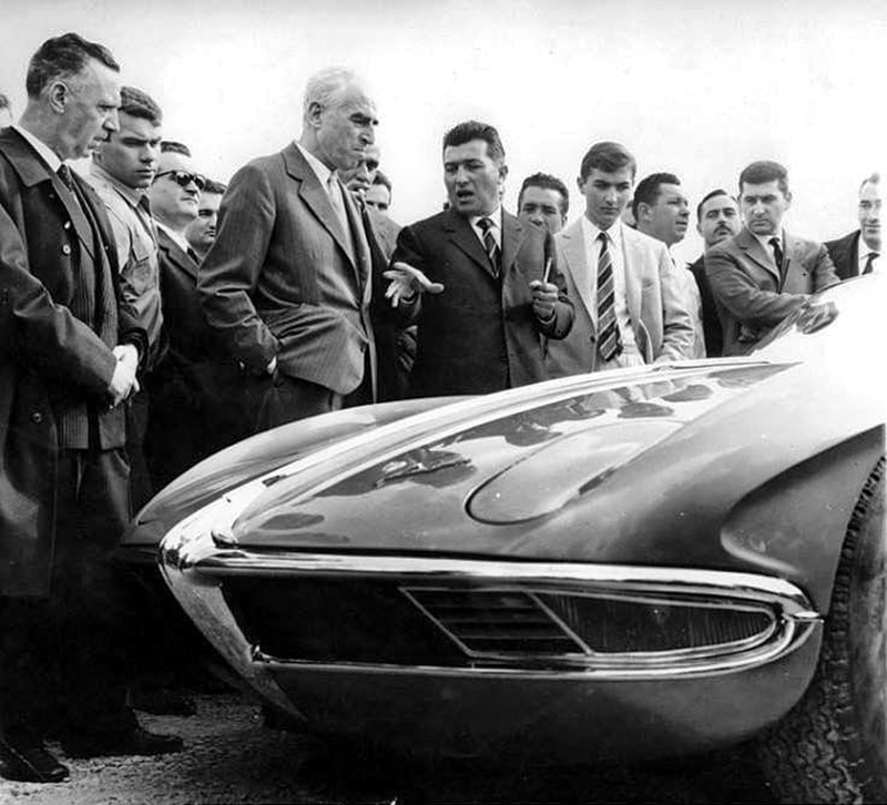 Several men wearing suits and ties stand next to the front of a car with sloped hood.