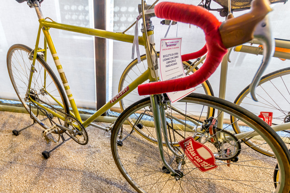A 3/4 profile view of a yellow racing bicycle with red wrapped handlebars sitting on a stand in a showroom
