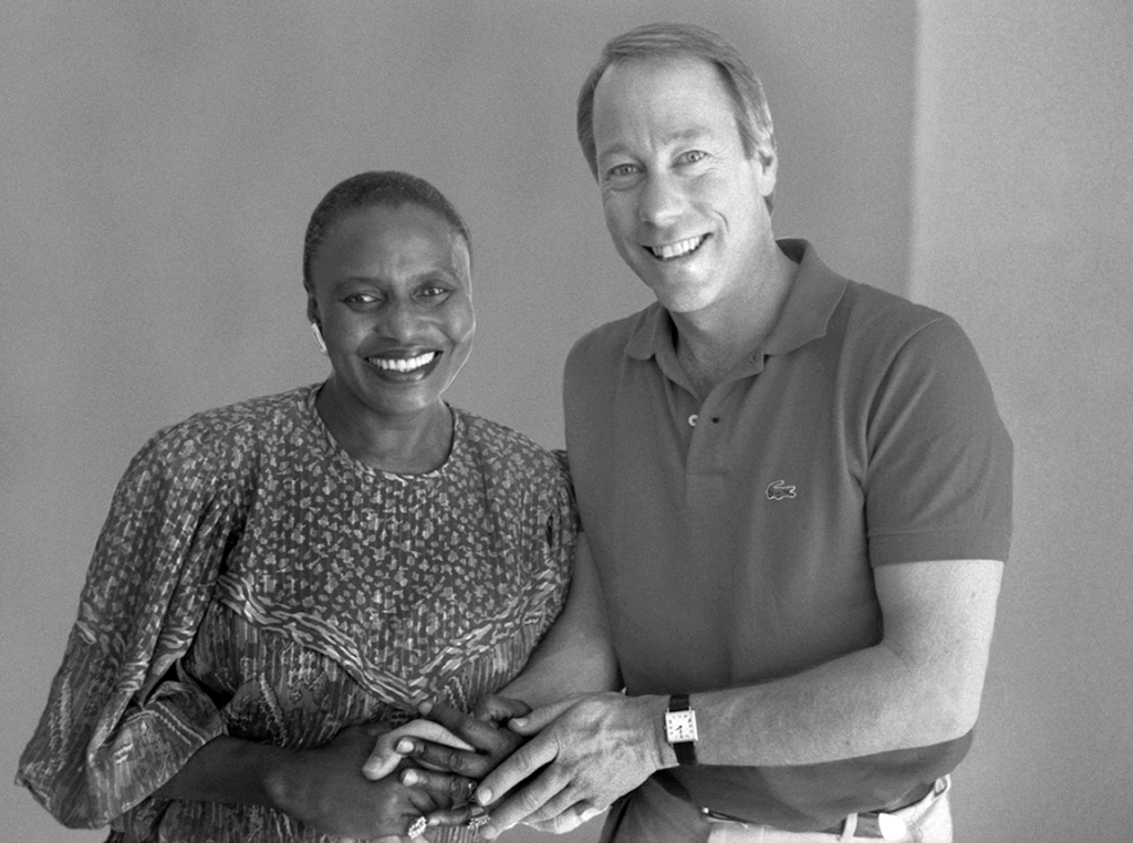 A woman with short hair wearing a patterned top clasps hands with a man in a polo shirt to her right, both smiling.