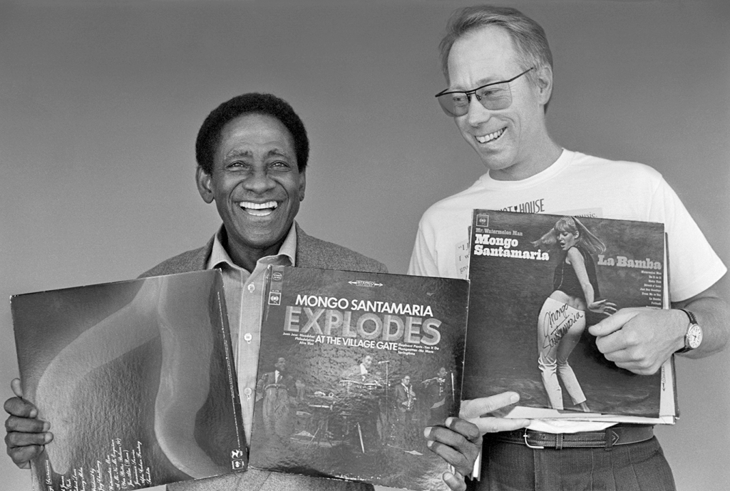 Two smiling men stand together. The man on the left holds two record albums, one titled “Mongo Santamaria Explodes at the Village Gate.” The man on the wears sunglasses and a casual shirt, holding a signed record album titled "Mongo Santamaria La Bamba."