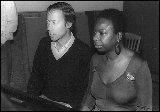 A man wearing a dark v-neck sweater over a white shirt sits at a piano to the left of a woman wearing long hoop earrings and a semi-sheer top with rhinestone decorations.