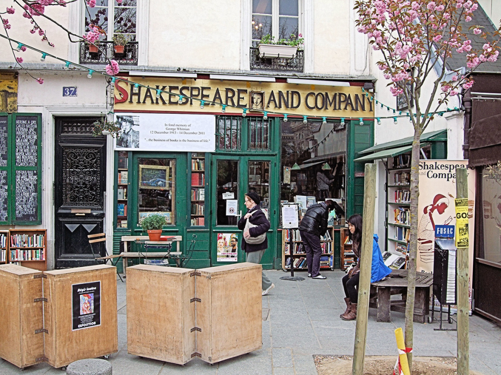 Storefront with large letters above the door and window that says "Shakespeare and Company." Many books can be seen both inside and outside the store on shelves.