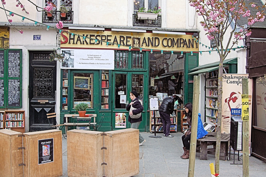 Storefront with large letters above the door and window that says "Shakespeare and Company." Many books can be seen both inside and outside the store on shelves.