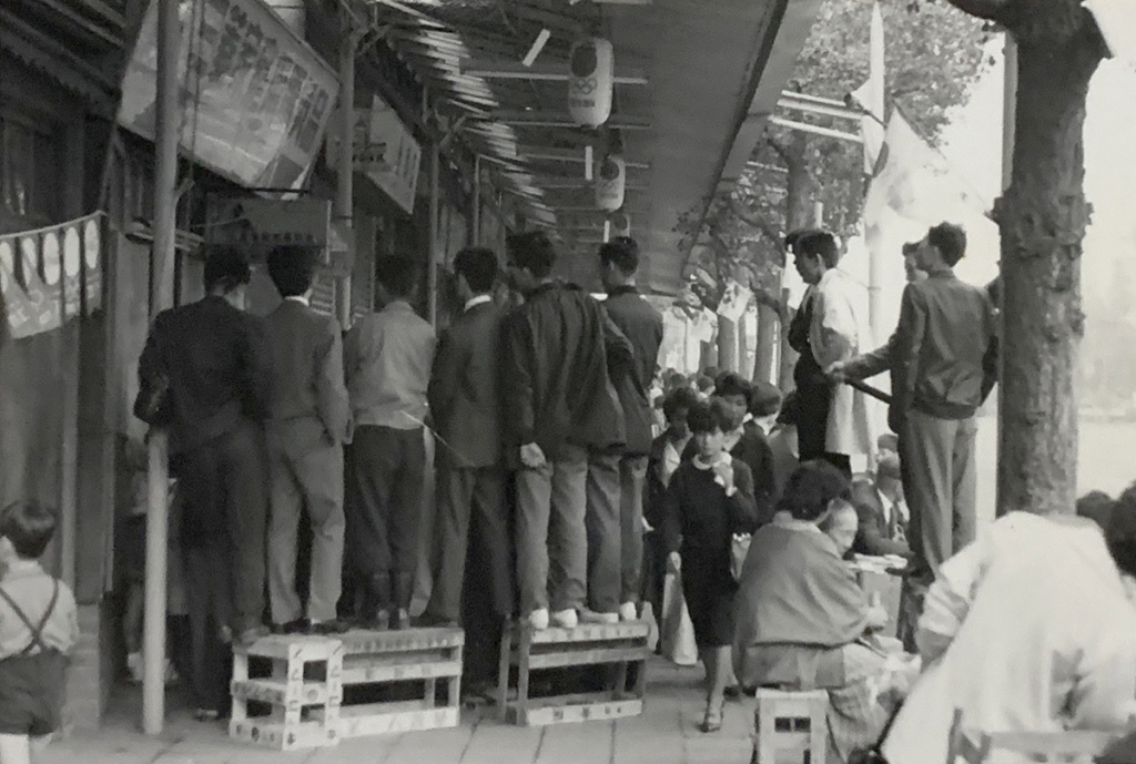 Black and white photograph of a crowded sidewalk showing several men in suits standing on crates with their backs to the camera. A woman wearing a dark-colored dress with a white collar walks around the standing men towards the camera.