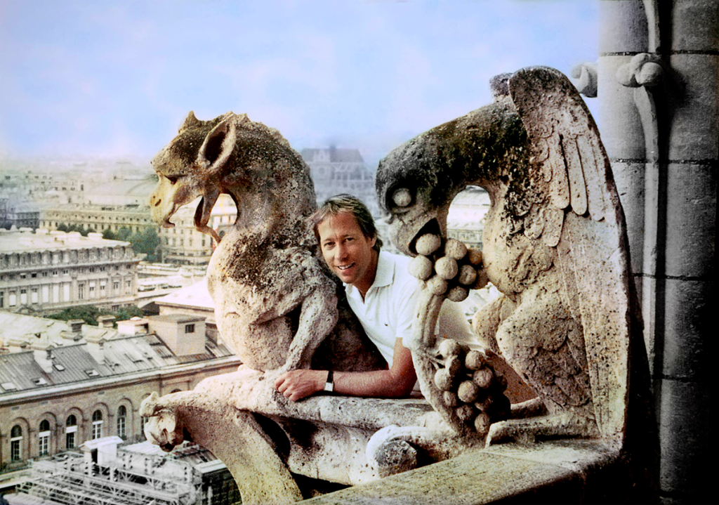 A smiling man poses between a pair of gargoyles with a city skyline in the background