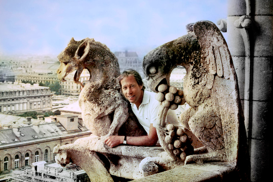 A smiling man poses between a pair of gargoyles with a city skyline in the background