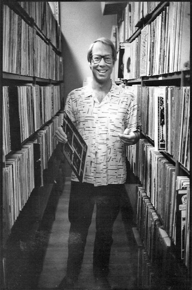 A smiling man wearing glasses and a patterned shirt stands in between two shelves holding record albums from floor to ceiling