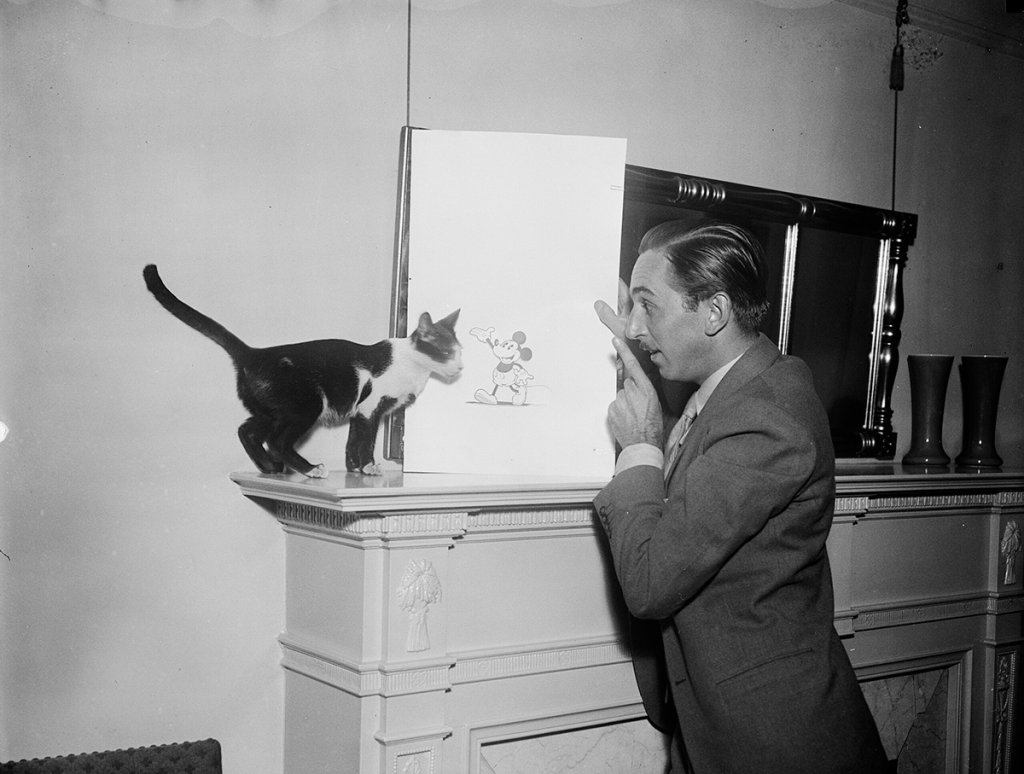 Well-dressed man stands in profile in front of a fireplace holding a drawing of a mouse propped on the mantle. He points a finger at the drawing and appears to be talking to a black and white cat standing on the other side of the drawing.