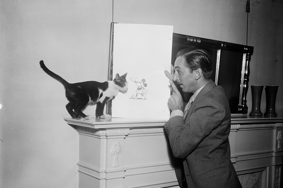 Well-dressed man stands in profile in front of a fireplace holding a drawing of a mouse propped on the mantle. He points a finger at the drawing and appears to be talking to a black and white cat standing on the other side of the drawing.