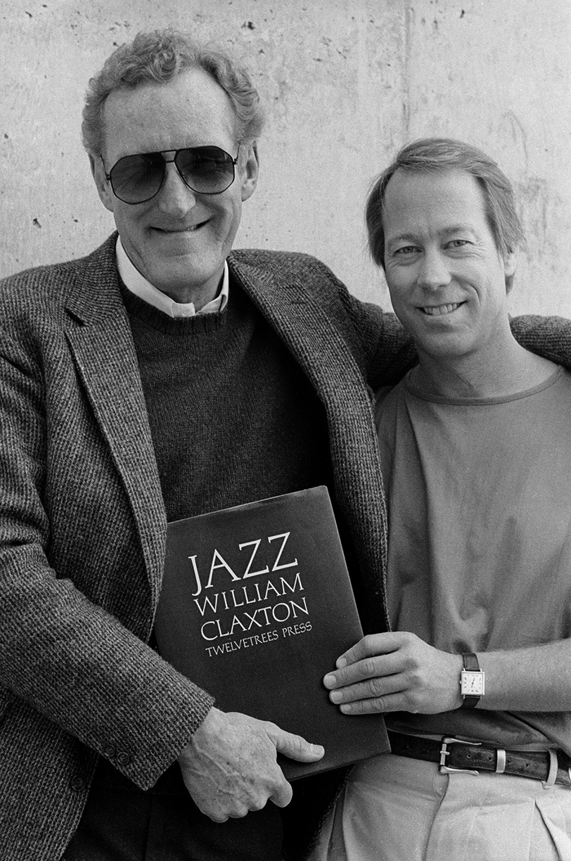 Two smiling men standing together. The man on the left wears sunglasses and a blazer and holds a book titled “Jazz William Claxton, Twelvetrees Press.” The man on the right rests his arm on his shoulder and wears a casual shirt and watch.