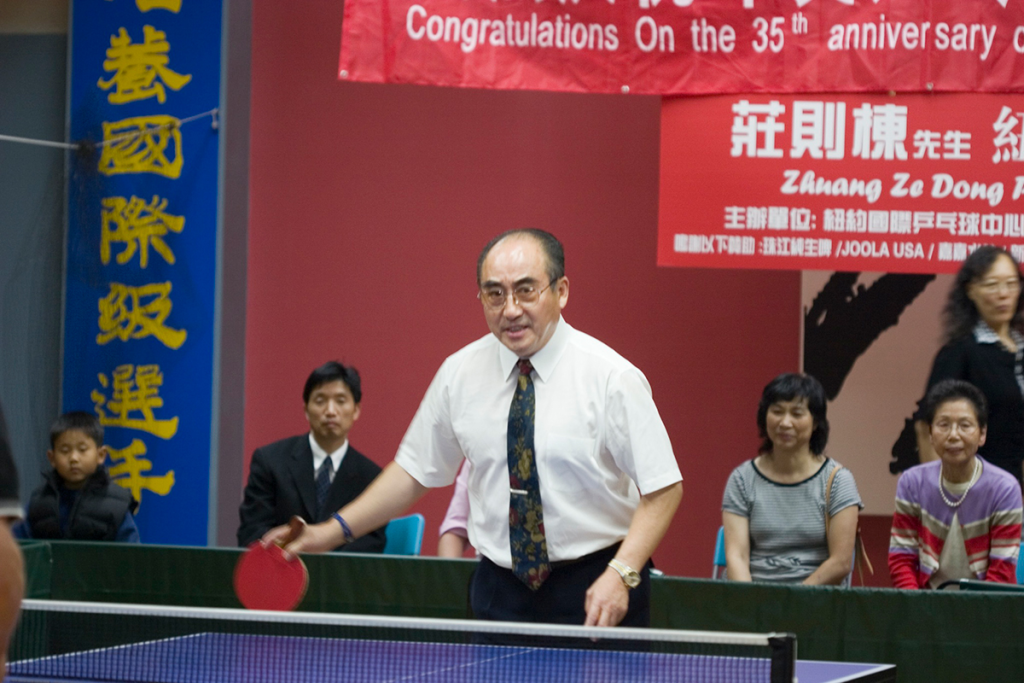 A middle-aged man with a receding hairline stands in front of a ping pong table. He is wearing glasses, short-sleeved shirt, and a tie. He holds a ping pong racket in his right hand.